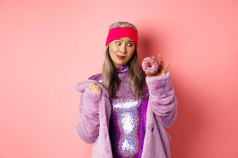 有趣的亚洲高级女人闪闪发光的迪斯科衣服人造皮毛外套诱惑美味的甜甜圈想要吃甜蜜的站粉红色的背景