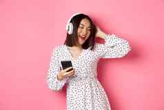 无忧无虑的亚洲女人有趣的跳舞听音乐耳机持有移动电话站衣服粉红色的背景