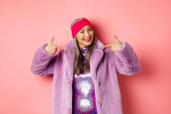时尚购物概念很酷的时尚的亚洲高级女人指出手指穿着时尚的紫色的人造革外套衣服微笑快乐粉红色的背景