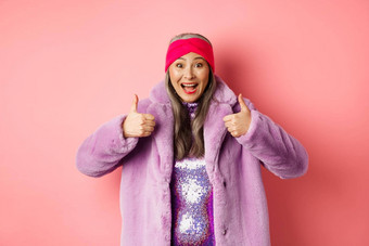时尚购物概念快乐的亚洲女人显示竖起大拇指批准推荐促销活动站时尚的<strong>紫色</strong>的皮毛<strong>外套</strong>粉红色的背景