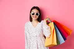 年轻的亚洲女人太阳镜购物持有袋购物中心商店微笑站粉红色的背景