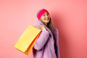 时尚的亚洲高级女人购物携带纸袋肩膀微笑快乐相机站粉红色的背景