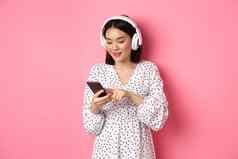 美丽的亚洲女人发短信消息智能手机听音乐耳机站粉红色的背景