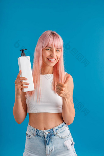 微笑女人光滑的粉红色的头发显示拇指持有泵瓶头发面具洗发水站孤立的蓝色的工作室背景