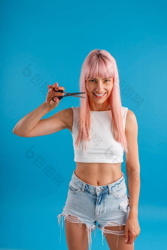 好玩的年轻的女人粉红色的头发微笑相机持有剪刀站孤立的蓝色的工作室背景