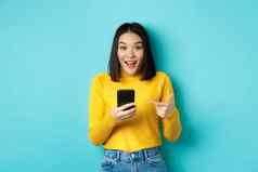 电子商务在线购物概念惊讶亚洲女人演示智能手机应用程序互联网折扣指出手指移动电话蓝色的背景
