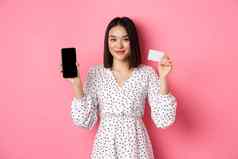 可爱的亚洲女人购物在线显示银行信贷卡移动屏幕微笑相机站粉红色的背景