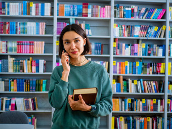 年轻的千禧女人会说话的细胞电话站公共图书馆背景书架特写镜头肖像女学生会说话的电话背景书架大学图书馆