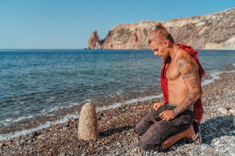 成熟的纹身男人。坐着海滩岩石祈祷准备好了战斗海背景肖像英俊的肌肉发达的古老的战士武术艺术概念