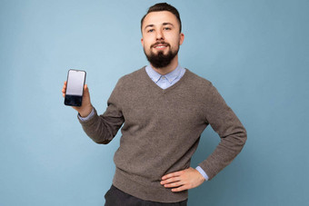 自信英俊的快乐很酷的年轻的浅黑肤色的女人不剃须的男人。beardwearing时尚的灰色毛衣蓝色的衬衫站孤立的粉红色的背景墙持有智能手机显示电话空屏幕显示相机