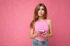 美丽的深思熟虑的年轻的金发女郎女人穿粉红色的前平衡孤立的粉红色的背景空空间持有手移动电话冲浪互联网在线一边怀疑