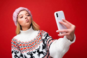 照片漂亮的积极的年轻的金发女郎女人穿温暖的针织他冬天温暖的毛衣站孤立的红色的背景移动电话采取自拍照片设备显示新加坡政府投资公司吻