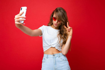 照片令人惊异的美丽的年轻的金发女郎女人持有移动电话采取自拍照片智能手机相机穿太阳镜日常时尚的装孤立的色彩斑斓的墙背景设备屏幕