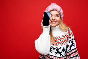 照片有吸引力的微笑年轻的金发女郎女人穿温暖的针织他冬天温暖的毛衣站孤立的红色的背景显示智能手机空屏幕断路一边