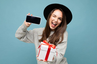有吸引力的快乐快乐的年轻的浅黑肤色的女人女人孤立的蓝色的背景墙穿时尚黑色的他休闲灰色毛衣持有礼物盒子显示移动电话屏幕显示模型相机眨眼
