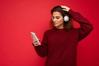 拍摄美丽的年轻的浅黑肤色的女人卷曲的女人穿黑暗<strong>红色</strong>的毛衣孤立的<strong>红色</strong>的<strong>背景墙</strong>穿白色无线耳机听音乐移动电话冲浪互联网智能手机显示