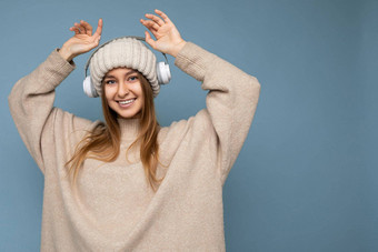 有吸引力的积极的微笑年轻的金发女郎女人穿米色冬天毛衣他孤立的蓝色的背景墙穿白色无线<strong>蓝牙耳机</strong>听音乐有趣的相机