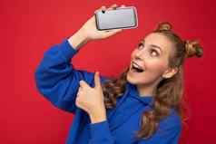 有吸引力的震惊了微笑年轻的金发女郎女人穿时尚的蓝色的连帽衫孤立的红色的背景复制空间持有智能手机显示电话手空屏幕显示指出gadjet开放口