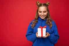 拍摄有吸引力的快乐微笑年轻的金发女郎女人孤立的红色的背景墙穿蓝色的时尚的连帽衫持有礼物盒子白色盒子红色的丝带
