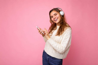 照片美丽的快乐的微笑年轻的女人穿时尚的休闲衣服孤立的背景墙持有移动电话穿白色蓝牙耳机听音乐有趣的相机