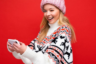 特写镜头照片眨眼美丽的微笑年轻的金发女郎女人穿温暖的针织他冬天温暖的毛衣站孤立的红色的背景玩游戏智能手机相机