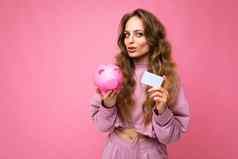 肖像照片快乐积极的年轻的美丽的有吸引力的女人波浪长金发女郎头发真诚的情绪穿时尚的粉红色的连帽衫孤立的粉红色的背景复制空间持有粉红色的小猪盒子信贷卡钱概念