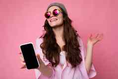 积极的微笑有吸引力的年轻的浅黑肤色的女人女人穿时尚的粉红色的衬衫灰色他色彩鲜艳的太阳镜孤立的粉红色的背景持有手显示移动电话空显示模型相机