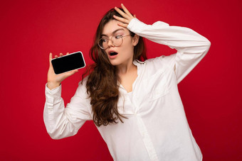 惊讶迷人的年轻的褐色女人穿白色衬衫光学眼镜孤立的红色的背景持有手显示移动电话空屏幕模型gadjet屏幕