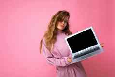 照片美丽的金发碧眼的卷曲的女人持有电脑移动PC空监控屏幕模拟复制空间穿粉红色的毛衣孤立的粉红色的墙背景