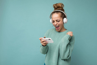 有吸引力的情感积极的年轻的女人穿蓝色的毛衣孤立的蓝色的背景穿白色蓝牙无线耳机听音乐移动电话玩在线游戏gadjet屏幕有趣的