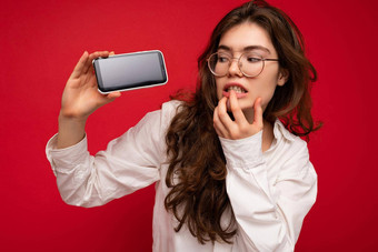 深思熟虑的有吸引力的年轻的褐色女人穿白色衬衫光学眼镜孤立的红色的背景持有手显示移动电话空屏幕模型gadjet屏幕有关