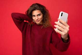 美丽的年轻的女人卷曲的头发穿黑暗红色的毛衣孤立的红色的背景墙持有聪明的电话电话屏幕采取自拍