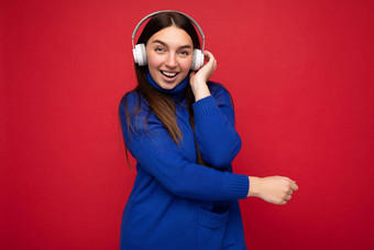 有吸引力的积极的年轻的浅黑肤色的女人女人穿蓝色的毛衣孤立的<strong>红色</strong>的<strong>背景墙</strong>穿白色蓝牙耳机听很酷的音乐有趣的相机