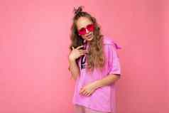 迷人的伤心年轻的金发女郎卷曲的女人孤立的粉红色的背景墙穿休闲粉红色的t恤时尚的色彩鲜艳的太阳镜相机