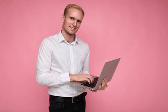 一边配置文件照片拍摄英俊的微笑快乐自信金发女郎男人。持有电脑移动PC打字键盘穿白色衬衫相机孤立的粉红色的背景
