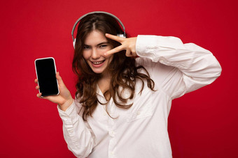 美丽的快乐微笑年轻的女人穿时尚的休闲装孤立的背景墙持有显示移动电话空显示模型穿白色蓝牙耳机听音乐有趣的相机