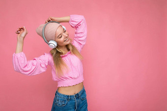 美丽的快乐微笑年轻的金发女郎女人穿粉红色的上衣粉红色的他孤立的粉红色的背景墙穿白色无线蓝牙<strong>耳机</strong>听很酷的音乐有趣的跳舞