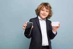 自信英俊的积极的快乐男孩卷曲的头发穿西装持有电话信贷卡显示移动屏幕相机孤立的蓝色的背景相机