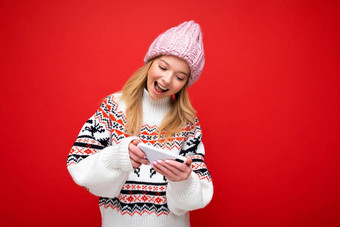 照片美丽的微笑年轻的金发女郎女人穿温暖的针织他冬天温暖的毛衣站孤立的红色的背景玩游戏智能手机设备显示
