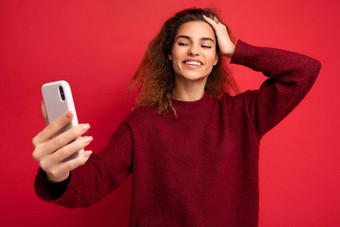 微笑快乐美丽的年轻的女人卷曲的头发穿黑暗红色的毛衣孤立的红色的背景墙持有聪明的电话电话屏幕采取自拍