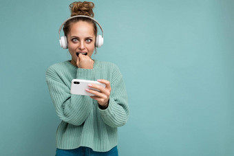 有吸引力的情感年轻的女人穿蓝色的毛衣孤立的蓝色的背景穿白色蓝牙无线耳机听音乐移动电话玩在线游戏相机有关