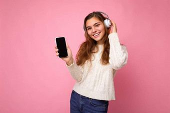 有吸引力的积极的微笑年轻的女人穿时尚的休闲装孤立的色彩鲜艳的背景墙持有显示移动电话空屏幕断路穿白色<strong>蓝牙耳机</strong>有趣的相机