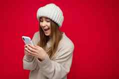 美丽的惊讶年轻的金发女郎女人穿休闲米色毛衣米色他孤立的红色的背景持有手移动电话沟通在线互联网gadjet显示