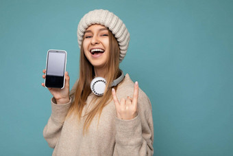 有吸引力的积极的微笑年轻的女人穿时尚的休闲装孤立的色彩鲜艳的背景墙持有显示移动电话空屏幕断路穿白色<strong>蓝牙耳机</strong>有趣的相机显示岩石卷手势标志
