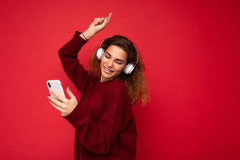 拍摄美丽的微笑年轻的浅黑肤色的女人卷曲的女人穿黑暗红色的毛衣孤立的红色的背景墙穿白色无线耳机听音乐移动电话冲浪互联网跳舞