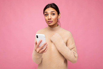 有吸引力的可爱的有关年轻的褐色女人穿米色温暖的毛衣站孤立的粉红色的背景冲浪互联网电话相机保持手心