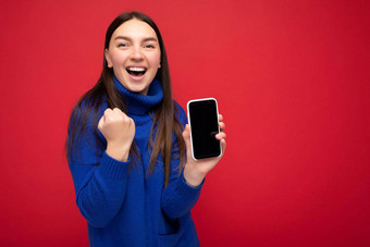 美丽的快乐年轻的浅黑肤色的女人女人穿休闲蓝色的毛衣孤立的红色的背景空空间持有手移动电话显示智能手机空屏幕模型相机