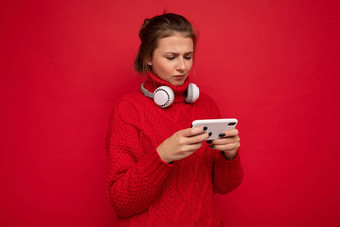 有吸引力的心烦意乱年轻的褐色女人穿红色的毛衣孤立的红色的背景墙持有移动电话冲浪互联网穿<strong>蓝牙耳机</strong>gadjet