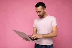 英俊的褐色男人。持有移动PC电脑打字键盘上网本监控t恤孤立的粉红色的背景