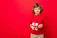 拍摄快乐积极的金发碧眼的卷曲的男孩站孤立的红色的背景墙穿红色的毛衣持有礼物盒子相机